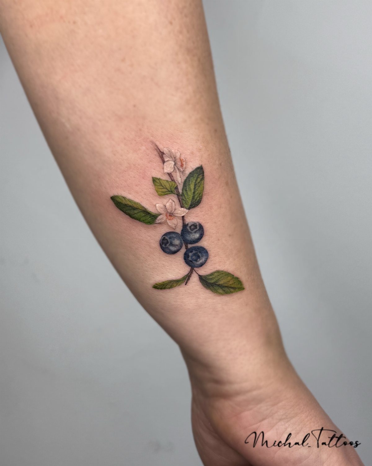 A realistic blueberry branch tattoo with green leaves and white flowers on a person's forearm.