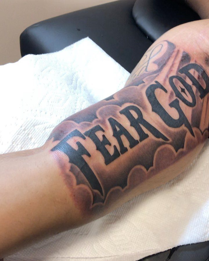 A large black and grey tattoo on an arm reading FEAR GOD against clouds.