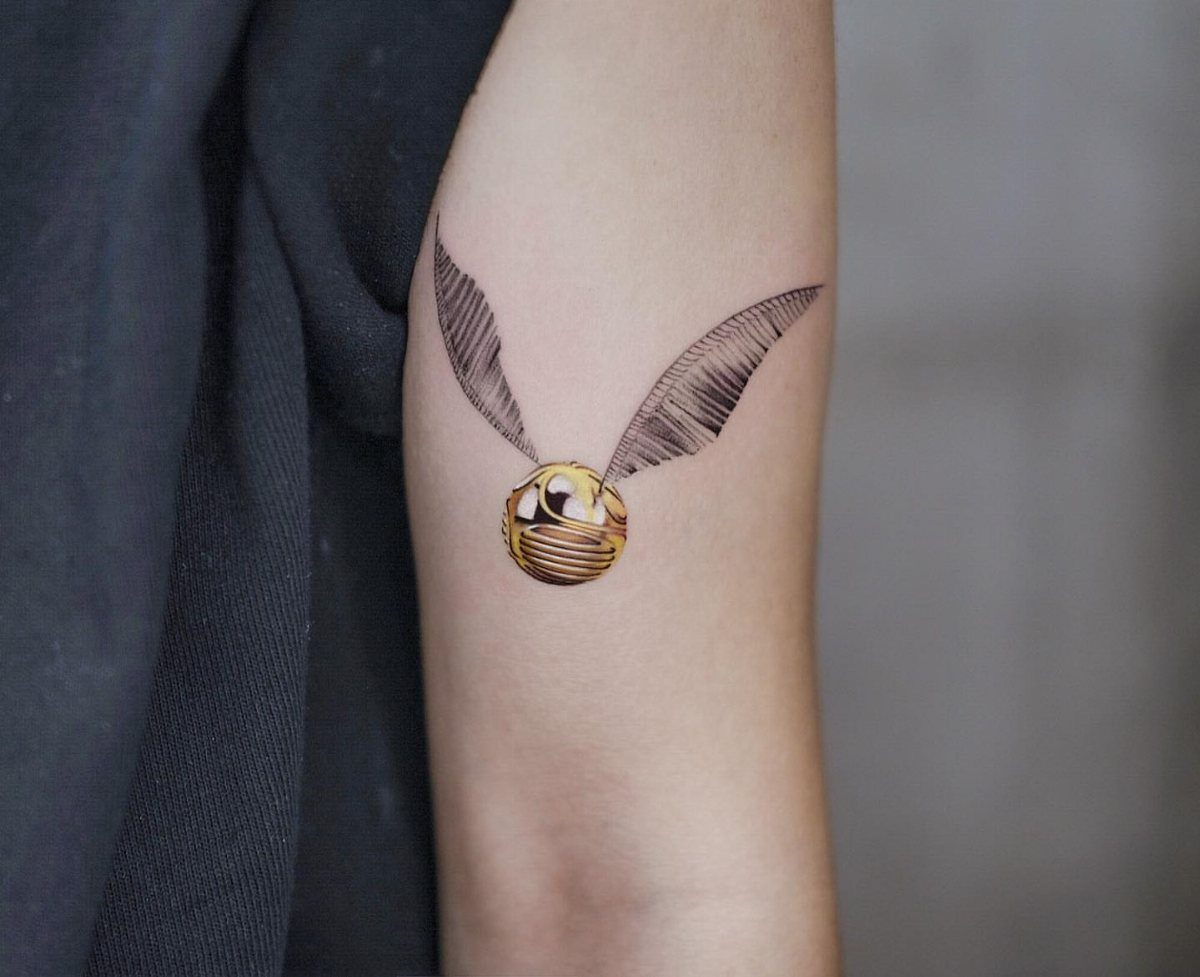 A detailed golden snitch tattoo with feathered wings on a person's arm.
