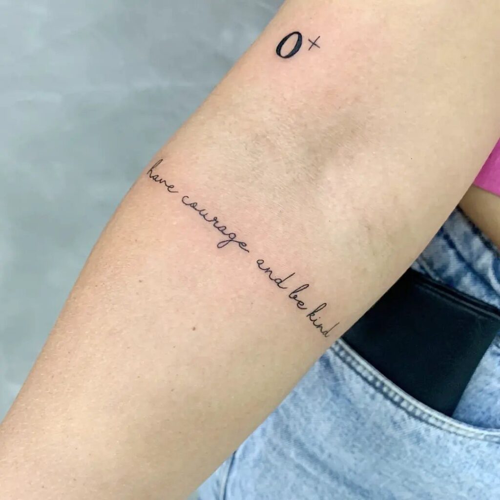 A forearm featuring a delicate script tattoo reading have courage and be kind.