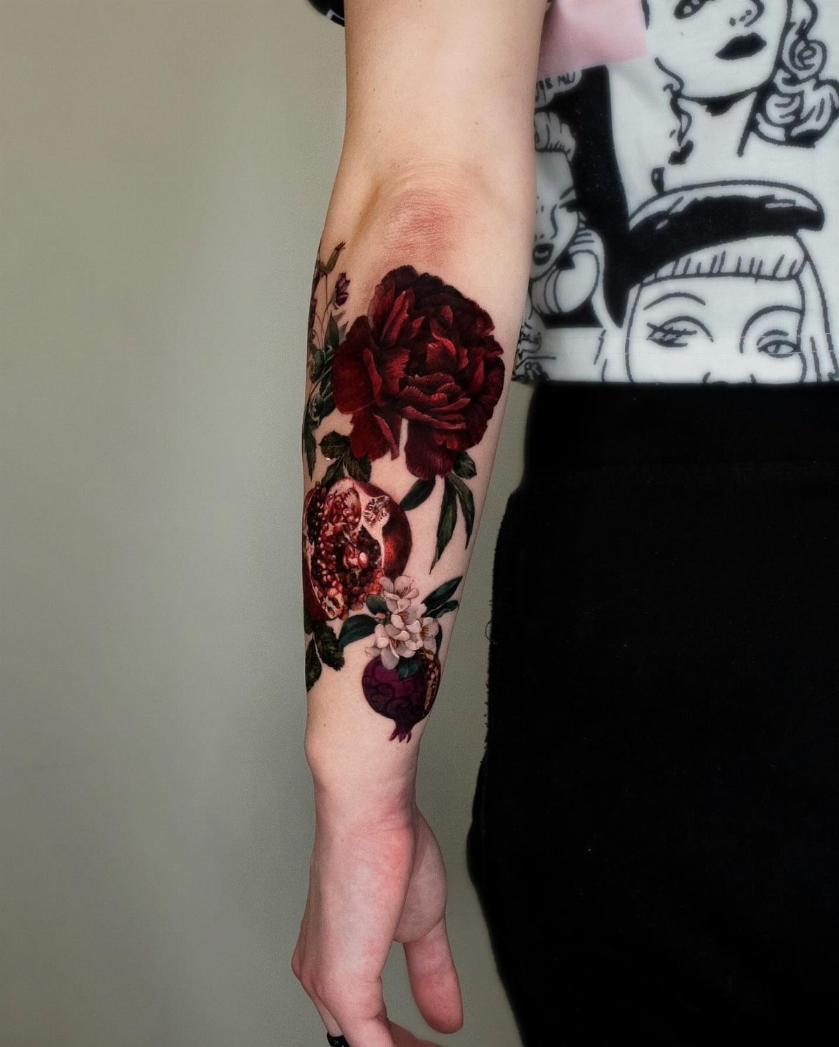 A detailed forearm tattoo of a red rose and a sliced pomegranate with green leaves.