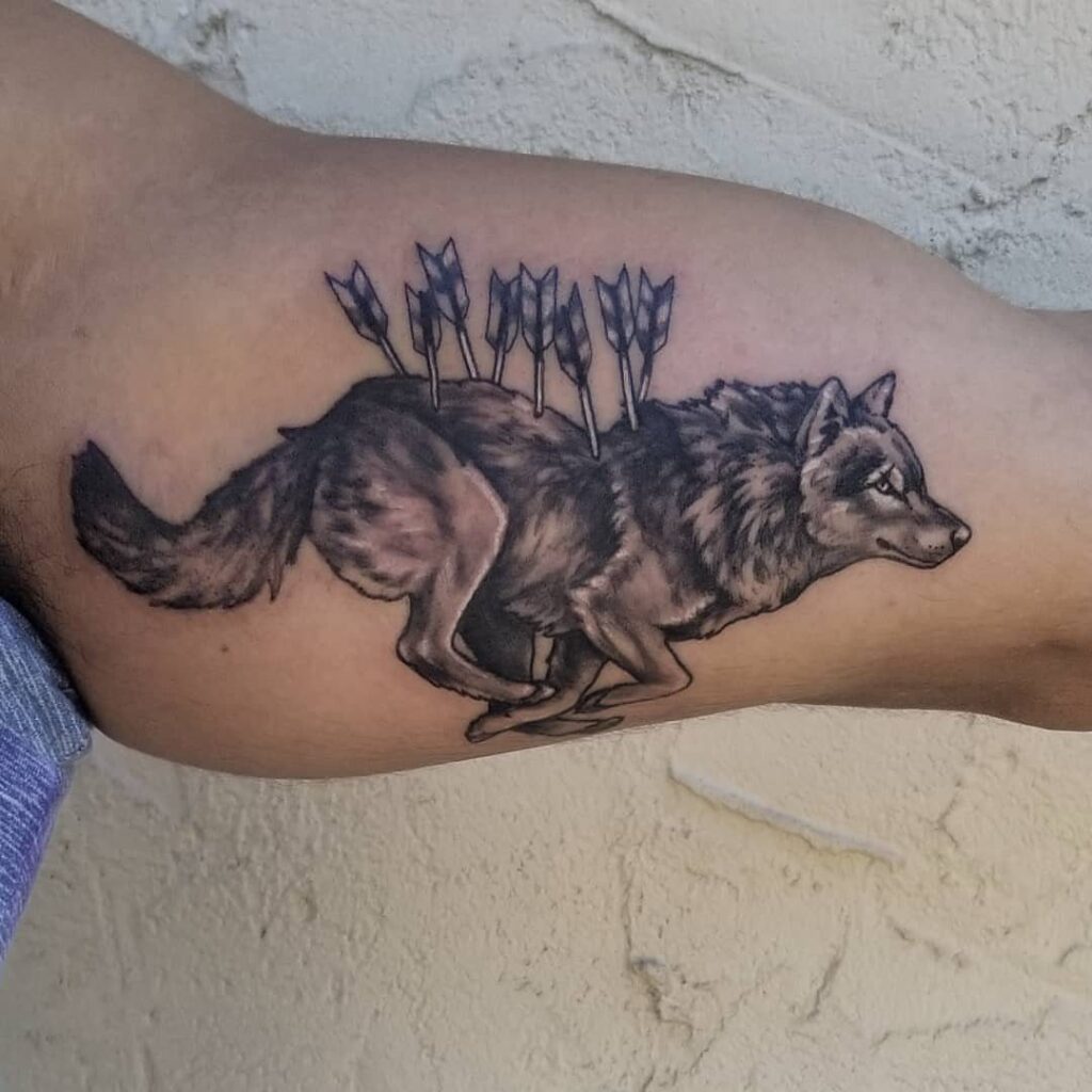 A black and grey tattoo of a running wolf with arrows in its back on an arm.