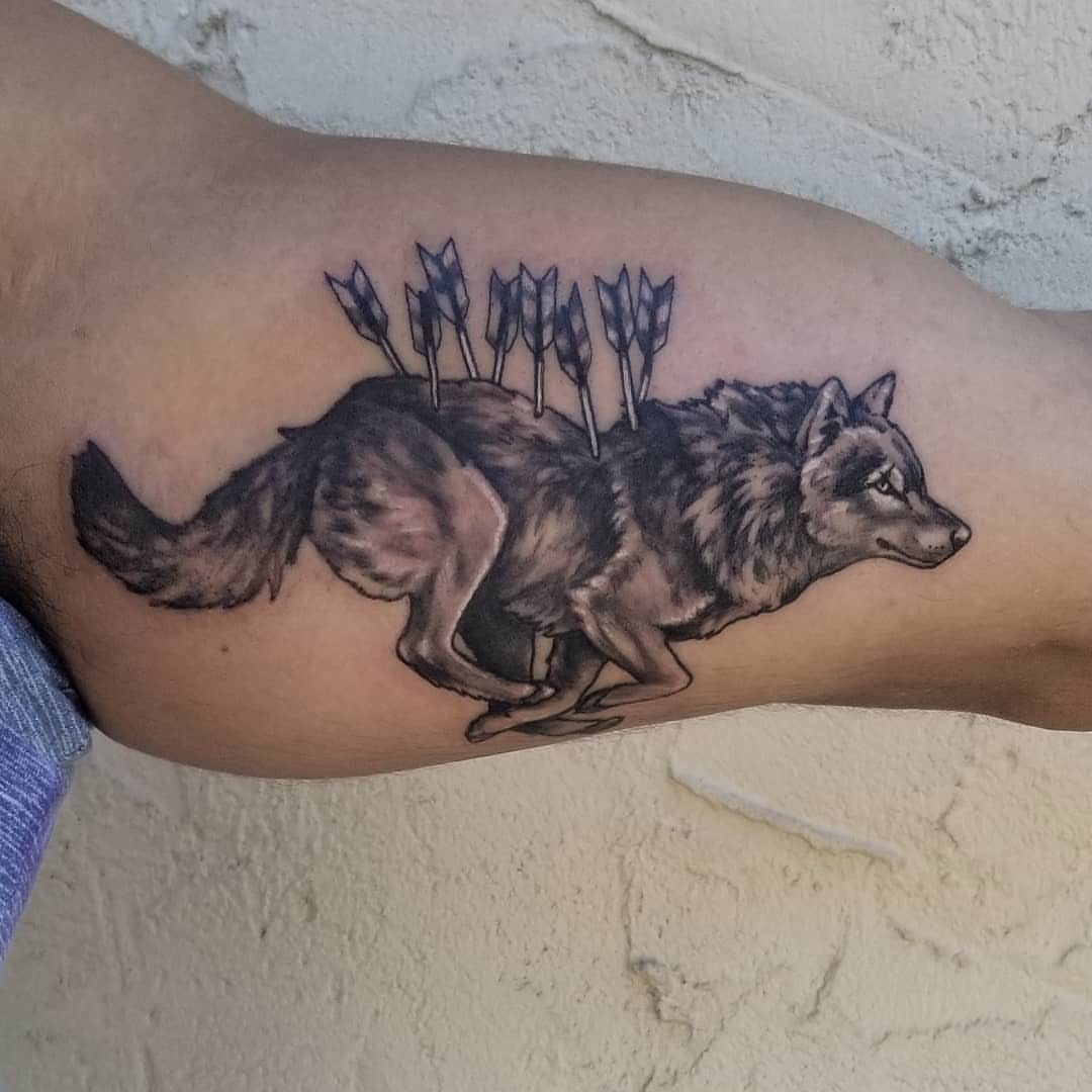 A black and grey tattoo of a running wolf with arrows in its back on an arm.