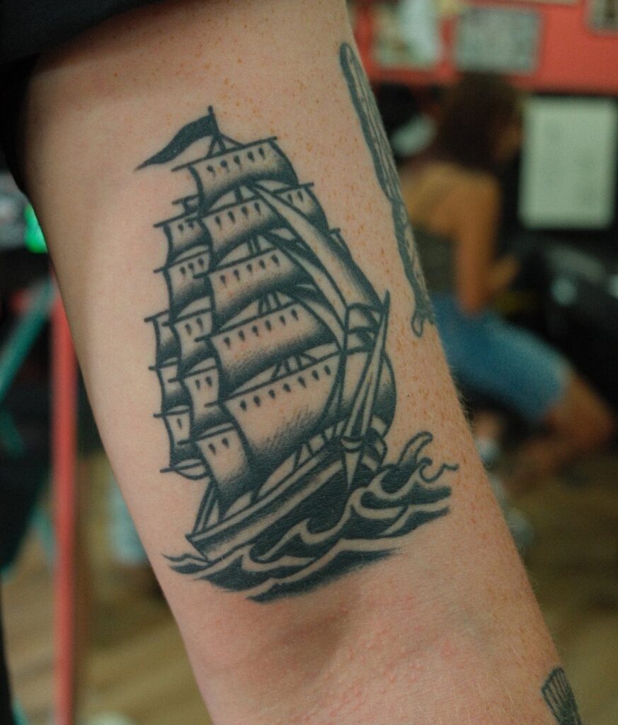 A black ink tattoo of a sailing ship on a person's forearm.