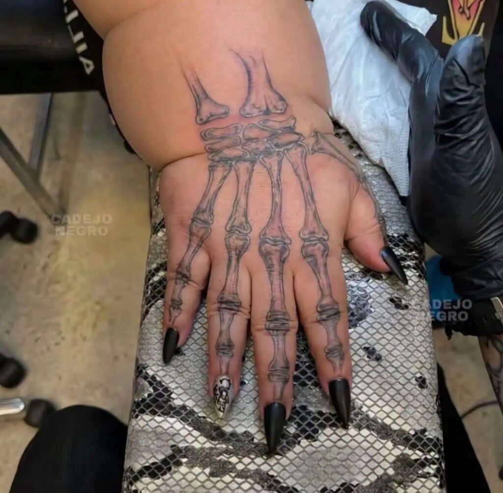 A realistic black and grey tattoo of anatomical hand bones on a person's hand.