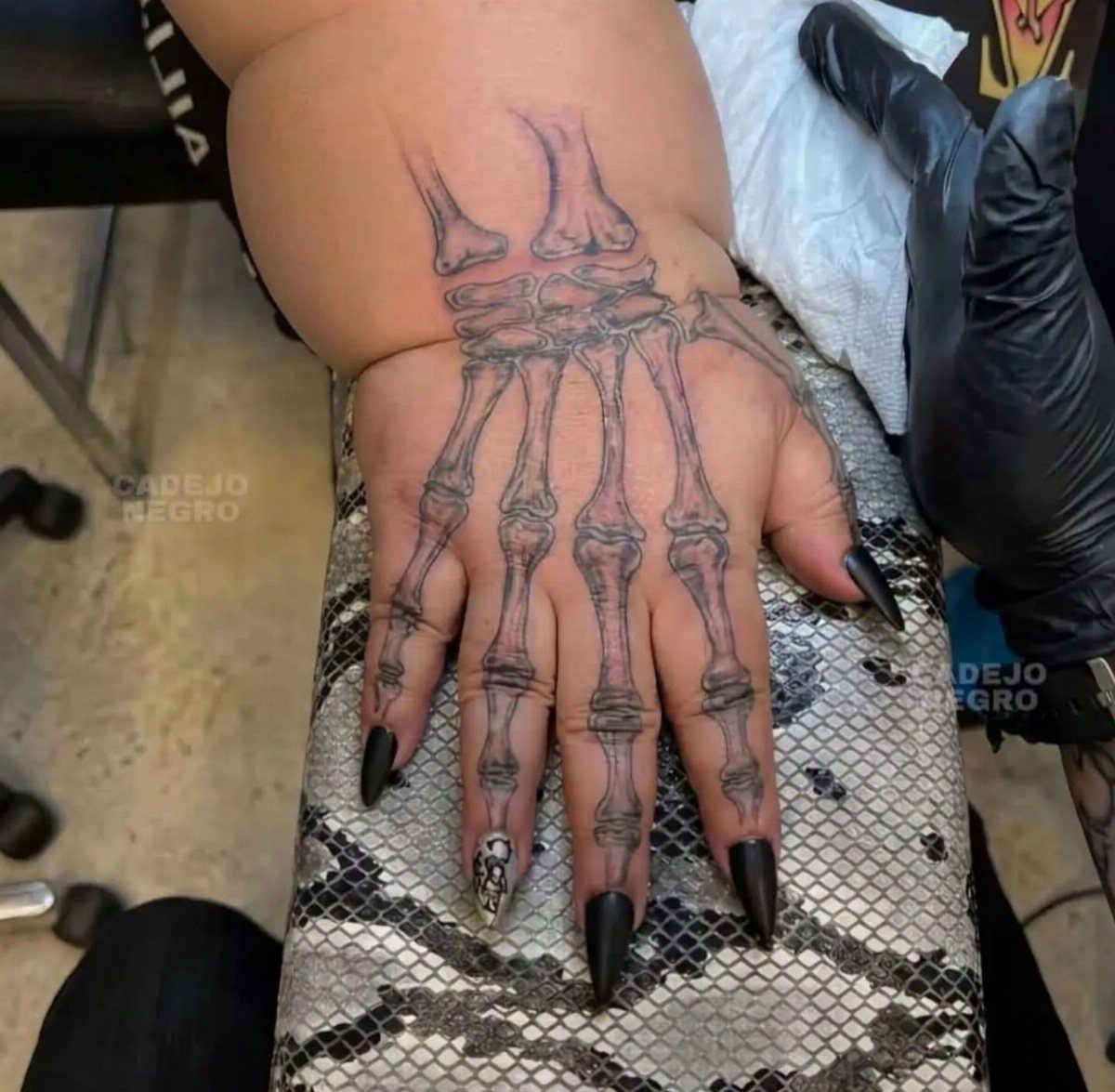 A realistic black and grey tattoo of anatomical hand bones on a person's hand.