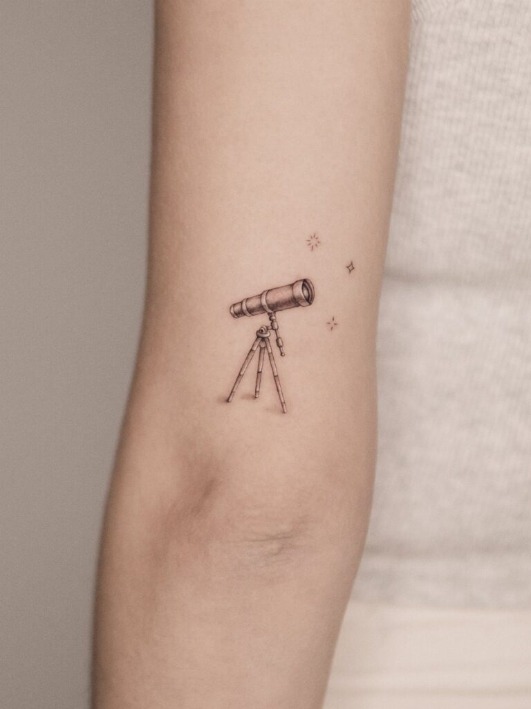 A small black ink telescope tattoo on a person's forearm with tiny stars.