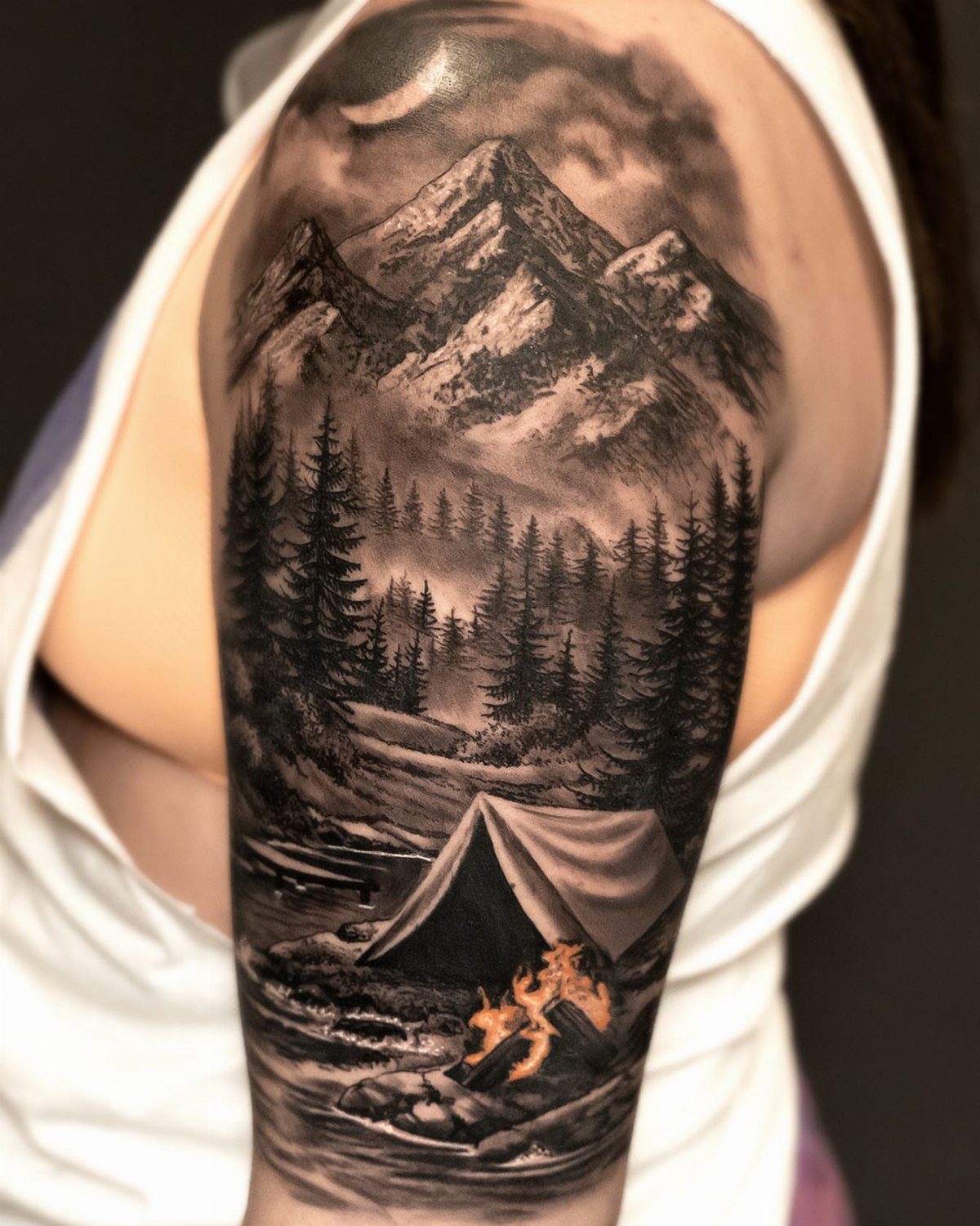 A black and grey mountain landscape tattoo featuring a tent and campfire on a person's arm.