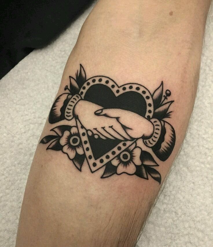 A black ink traditional handshake tattoo on a person's forearm featuring a heart and flowers.